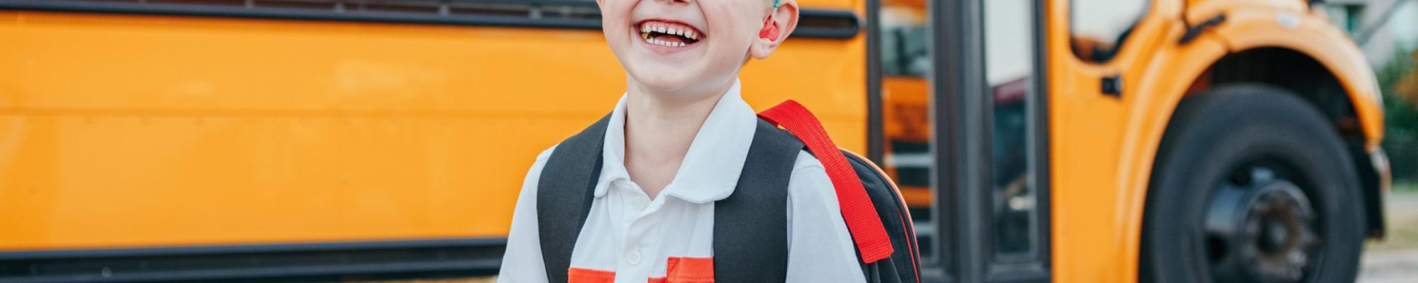Happy boy with backpack and apple in front of school bus.